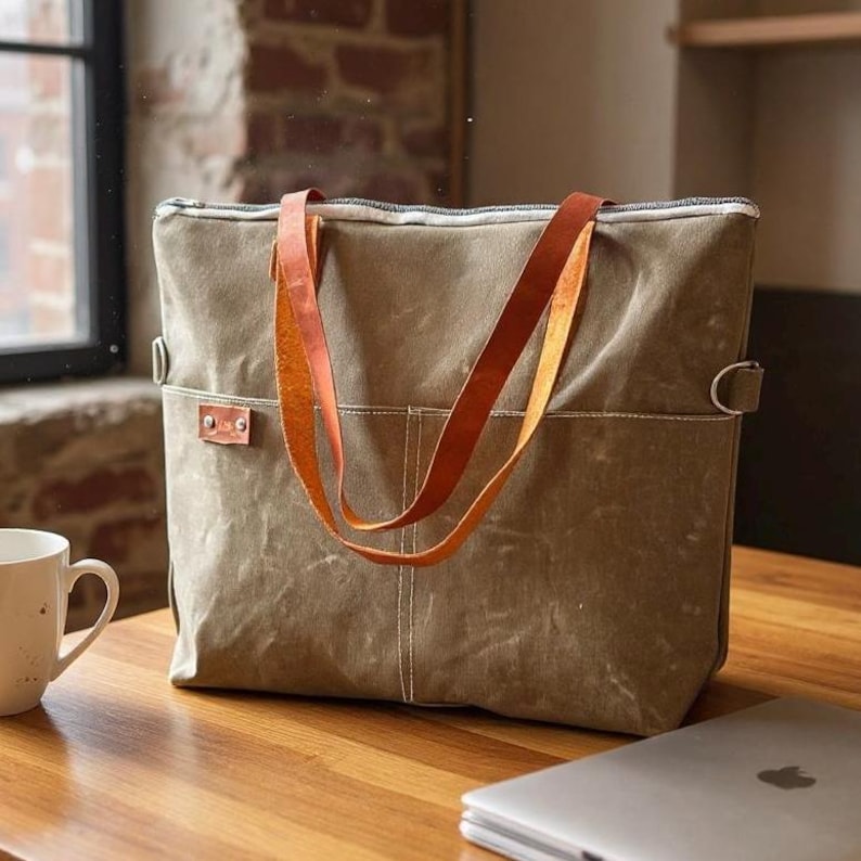K&ouml;nnte beinhalten: Eine gro&szlig;e, olivgr&uuml;ne Canvas-Tragetasche mit braunen Ledergriffen. Die Tasche hat einen Rei&szlig;verschluss und zwei Au&szlig;entaschen. Die Griffe sind mit Metallbeschl&auml;gen befestigt. Die Tasche steht auf einem Holztisch.