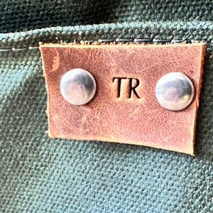 K&ouml;nnte beinhalten: Nahaufnahme eines braunen Lederetiketts mit den gepr&auml;gten Initialen "TR". Zwei silberne Nieten befestigen das Etikett an einem strukturierten, dunkelgr&uuml;nen Stoff. Das Etikett ist rechteckig mit abgerundeten Ecken.