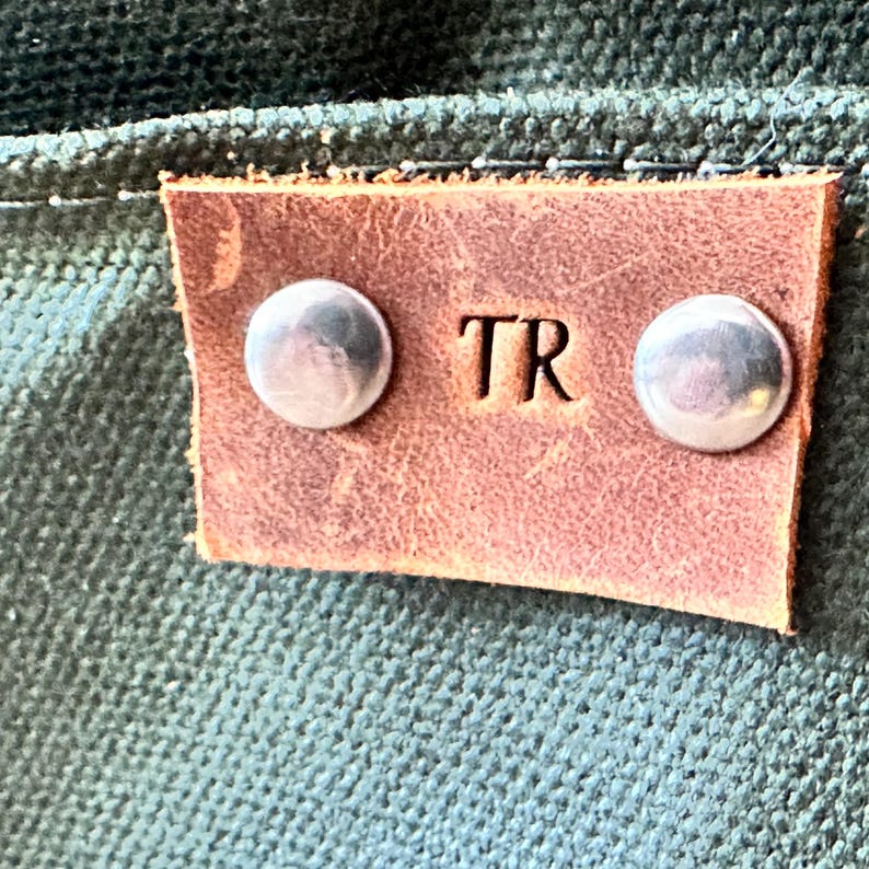 K&ouml;nnte beinhalten: Nahaufnahme eines braunen Lederetiketts mit den gepr&auml;gten Initialen "TR". Zwei silberne Nieten befestigen das Etikett an einem strukturierten, dunkelgr&uuml;nen Stoff. Das Etikett ist rechteckig mit abgerundeten Ecken.