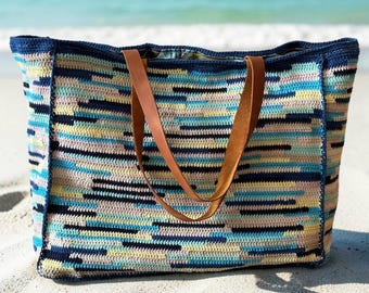 May include: A rectangular crochet tote bag featuring a horizontal striped pattern in shades of navy blue, light blue, pale yellow, and soft pink. The bag includes two brown leather handles and a dark blue crocheted trim along the top edge.