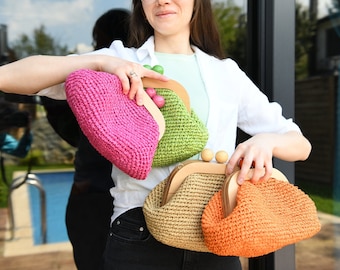 May include: Four woven clutch purses in shades of pink, green, tan, and orange. Each purse has a wooden frame and a clasp with wooden beads. The purses are held up in front of a person.