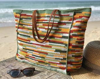 May include: A large, colorful crochet tote bag with brown leather handles. The bag features a striped pattern in shades of green, yellow, red, and white. Tortoiseshell sunglasses and a straw hat are next to the bag on a wooden surface, with the ocean in the background.