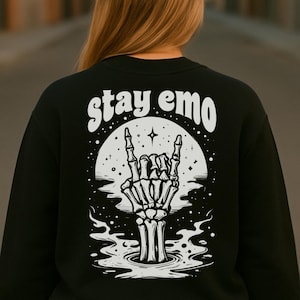 Può includere: Felpa nera con la scritta "stay emo" sopra una grafica bianca di una mano scheletrica che fa il gesto del rock and roll. La grafica è ambientata su uno sfondo di luna e acqua.