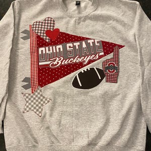 May include: A gray sweatshirt with a decorative design featuring the words "OHIO STATE Buckeyes" on a red pennant. The design includes a football, a foam finger, hearts, and stars. The sweatshirt is a crew neck style.