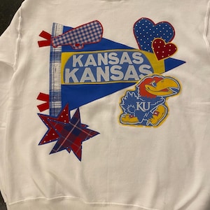 Pode incluir: Camisola branca com desenhos aplicados com o tema Kansas Jayhawks. Apresenta uma flâmula azul com "KANSAS" em amarelo e branco, um Jayhawk de desenho animado e detalhes xadrez e bolinhas. Uma peça de vestuário confortável com tema universitário.