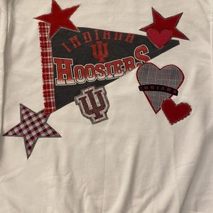 以下が含まれることがあります： 赤と白の文字で「インディアナ・フーシャーズ」と書かれたペナントデザインの白いスウェットシャツ。デザインには赤い星とハート、インディアナ大学のロゴが含まれています。