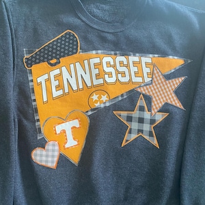 Puede incluir: Sudadera gris oscuro con la palabra "TENNESSEE" en letras blancas sobre un banderín naranja. La sudadera presenta estrellas y corazones naranjas y grises, y un diseño de megáfono.