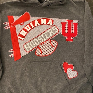 May include: A grey hooded sweatshirt with a front pocket and a drawstring hood. The sweatshirt features appliqued red and white plaid designs, including the word "INDIANA" and "HOOSIERS", a megaphone, a football, and two hearts.