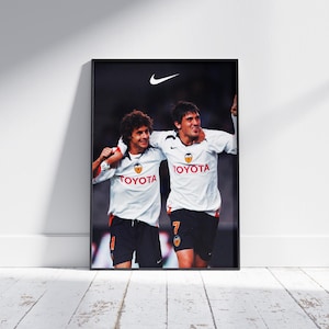 Può includere: Poster incorniciato con due calciatori in maglia bianca con il logo Toyota. I giocatori si abbracciano, festeggiando una vittoria. Il logo Nike è visibile sopra. Il poster è su un muro bianco.