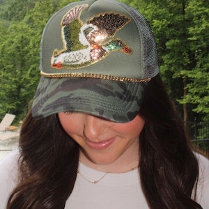 May include: A green camouflage trucker hat with a beaded duck design. The hat features a gold chain detail along the brim and a mesh back. The duck design includes shades of green, white, orange, and gold.