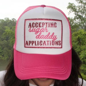 May include: A pink and white trucker hat with the text "ACCEPTING sugar daddy APPLICATIONS" in a glittery, pink font. The hat has a pink brim and a white front panel with a pink mesh back.