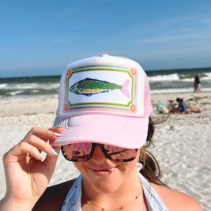 May include: A light pink trucker hat featuring a white front panel with a rectangular border and a graphic illustration of a green and yellow fish with a pink tail. The hat has a mesh back and a solid pink brim, designed with a structured crown.