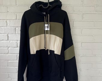 May include: A black hooded sweatshirt featuring a color-blocked design with a horizontal olive green panel across the chest and a beige panel below it. The hoodie includes a front kangaroo pocket, long sleeves with olive green accents, and a tag.