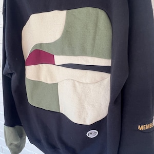 Könnte beinhalten: Ein schwarzer Kapuzenpullover mit einem großen, zentralen Aufnäher mit abstrakten, organischen Formen in Olivgrün, Creme und Dunkelburgunder. Das Kleidungsstück enthält ein kleines kreisförmiges Logo-Emblem, ein grünes Ärmelbündchen und das Wort MEMBE auf dem Ärmel.