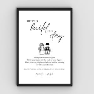 May include: Black framed wedding sign with the text "HELP US BUILD OUR DAY" in elegant script. Below the text are illustrations of Lego figures of a bride and groom. The sign includes instructions and names.