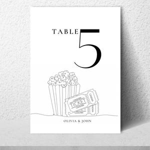 May include: A white rectangular table number card featuring the black text TABLE 5. Below the number is a black line drawing of a popcorn bucket and two cinema tickets. The names OLIVIA & JOHN are printed in black at the bottom of the card.