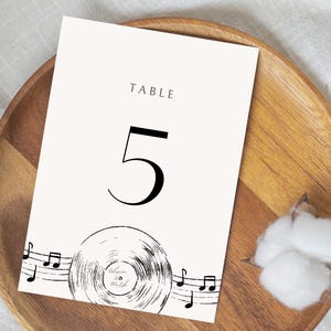May include: A cream-colored table number card with the number 5 in a bold black font. The card features the word "TABLE" at the top and a vinyl record illustration with musical notes at the bottom. The card is on a wooden plate.