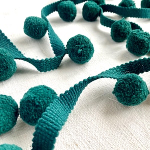 以下が含まれることがあります： ターコイズ色のポンポン飾りのクローズアップ。このトリムは、織られたターコイズ色のリボンに小さな丸いふわふわのポンポンが付いています。リボンはスカラップエッジです。このトリムは、おそらく工芸品や衣類やアクセサリーの装飾に使用されます。