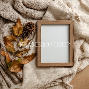 以下が含まれることがあります： ベージュ色のニットブランケットの上に、「FRAME MOCKUP」の文字が入った木製フレームのモックアップが置かれています。秋の葉と松ぼっくりが季節感を添えています。フレームは空で、デザインの準備ができています。
