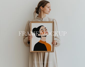 Op de afbeelding: Een vrouw houdt een ingelijst illustraties van een vrouw met gesloten ogen en oranje en zwarte kleding vast. De lijst is goudkleurig en de illustratie is op een witte achtergrond. De tekst "FRAME MOCKUP" is in witte letters op de afbeelding geschreven.