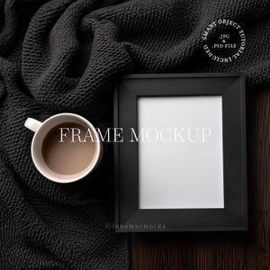May include: A black picture frame mockup with a white interior, next to a white mug filled with coffee. The frame is on a dark wooden surface, with a dark gray knitted blanket in the background. The text "FRAME MOCKUP" is displayed.