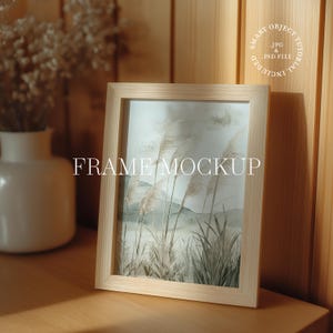May include: A light-colored wooden picture frame with a landscape print featuring tall grasses and mountains. The text "FRAME MOCKUP" is displayed in the center of the frame. The frame is on a wooden surface.
