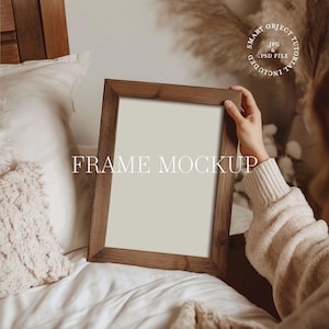 May include: A rectangular wooden frame mockup with the text "FRAME MOCKUP" in white. The frame is held by a person wearing a beige sweater. The background includes a bed with white bedding and decorative elements.