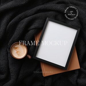 May include: A black picture frame mockup with a white interior, resting on a brown book. A cup of coffee sits nearby on a dark gray knitted blanket. The text "FRAME MOCKUP" is displayed in white.