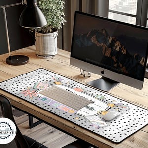 May include: A rectangular desk mat with a white background and black polka dots. The mat features a keyboard area with the words "BE KIND" and floral and pastel graphics. A computer mouse and keyboard are on the mat.