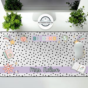 Op de afbeelding: Een rechthoekige bureaumat met een witte achtergrond en zwarte stippen. De mat heeft de woorden "BE KIND" in pastelkleuren, samen met illustraties van een ster, een boek, een bloem en een perzik. De naam "Mrs. Nelson" staat op een lavendelkleurige banner.