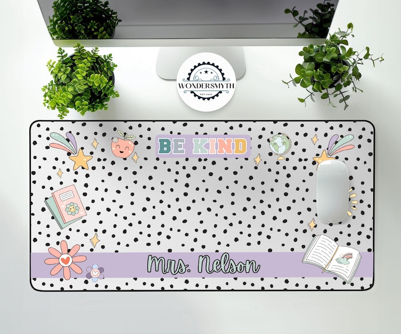 Op de afbeelding: Een rechthoekige bureaumat met een witte achtergrond en zwarte stippen. De mat heeft de woorden "BE KIND" in pastelkleuren, samen met illustraties van een ster, een boek, een bloem en een perzik. De naam "Mrs. Nelson" staat op een lavendelkleurige banner.