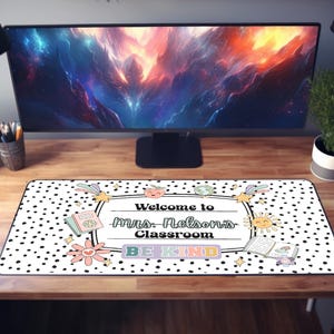 May include: A rectangular desk mat with a black and white polka dot pattern. The mat features the text "Welcome to Mrs. Nelson's Classroom" and "BE KIND" in a colorful, playful design. Decorative elements include flowers, books, and a sun.