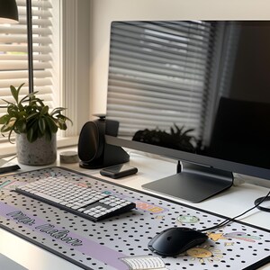 Op de afbeelding: Een moderne thuiskantooropstelling met een grote computermonitor, draadloos toetsenbord en muis op een bureau. Een decoratieve muismat met een stippenpatroon en de tekst "Mrs. Mason" is zichtbaar. Een potplant en luidsprekers zijn ook aanwezig.