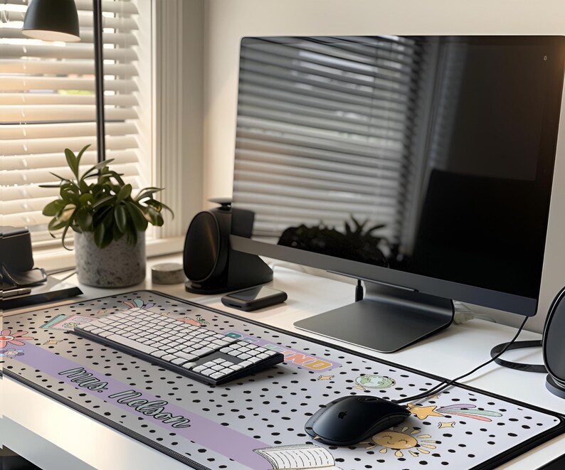 Op de afbeelding: Een moderne thuiskantooropstelling met een grote computermonitor, draadloos toetsenbord en muis op een bureau. Een decoratieve muismat met een stippenpatroon en de tekst "Mrs. Mason" is zichtbaar. Een potplant en luidsprekers zijn ook aanwezig.