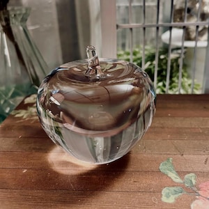 May include: A clear glass apple-shaped decorative object with a stem. The apple is sitting on a wooden surface with a floral design. The glass reflects the surrounding environment.