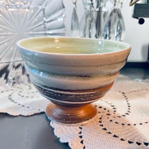 May include: A handcrafted ceramic bowl with a pedestal base. The bowl features a layered design with shades of beige, green, and brown. The interior is a light yellow color. The bowl is sitting on a white lace doily.