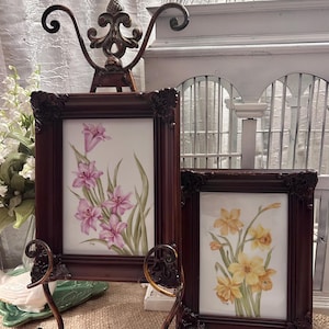 May include: Two framed floral paintings with ornate dark brown frames. One painting features pink lilies, the other yellow daffodils. The frames are displayed on decorative metal easels, with a white birdcage in the background.