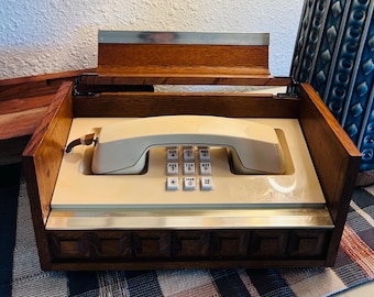May include: A retro cream-coloured telephone with a push-button keypad, housed in a wooden box with a hinged lid. The box has a dark wood finish with decorative trim and a metal accent. The phone is a vintage design, likely from the mid-20th century.