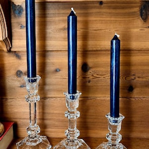 May include: Three clear crystal candle holders with navy blue taper candles. The candle holders have a faceted design and sit on a wooden surface. The candles are unlit and have white wicks.