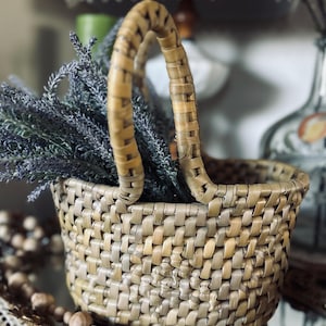 May include: A round woven basket with a single arched handle, constructed from light tan and beige natural fibers in a tight, interlocking pattern. The basket is filled with a cluster of dried purple lavender sprigs that extend upward.