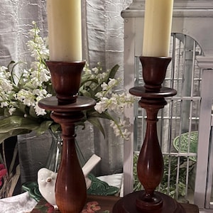 May include: Two antique wooden candle holders with cream-colored candles. The candle holders are dark brown with a turned design and a round base. The candles are tall and slender. The background includes white flowers and a decorative bird.