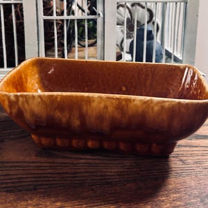 May include: A rectangular, brown ceramic planter with a glossy finish. The planter has a slightly flared shape and a decorative beaded base. The interior is a darker brown, and the exterior has a mottled, glazed appearance. The planter is designed for indoor or outdoor use.