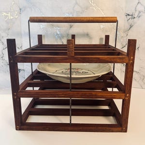 May include: A dark brown wooden tiered carrier with a cylindrical handle and metal support rods. Inside the frame sits a white ceramic pie dish featuring a graphic of cherries and the text "Cherry Pie" printed in black on the center surface.