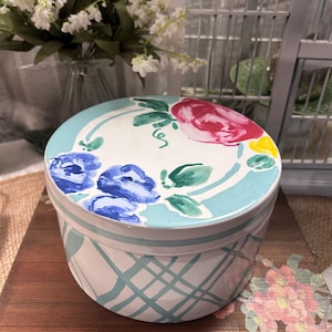 May include: A round, ceramic container with a lid. The lid is decorated with blue, pink, and yellow floral designs on a light blue background. The container's body features a white base with a light blue crisscross pattern.