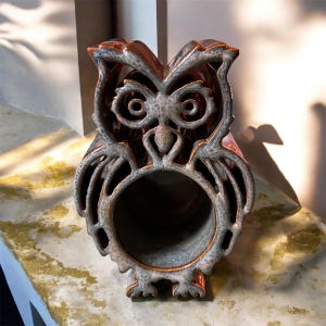 May include: A ceramic owl figurine with a hollow center. The owl has a detailed design with openwork wings and a textured, weathered appearance. The color palette includes shades of brown, gray, and a hint of red.