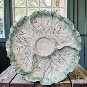 May include: A large, ceramic serving platter in the shape of a cabbage leaf. The platter is primarily light green with darker green edges and a central dip bowl. The leaf design features detailed veining, adding to its realistic appearance. This decorative piece is ideal for serving appetizers or snacks.