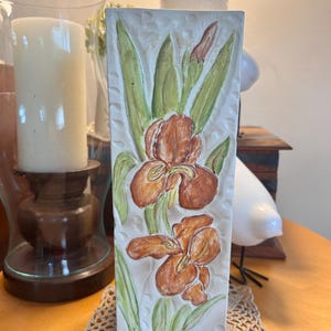May include: A rectangular ceramic vase with a white base featuring a raised, hand-painted relief of two brown iris flowers and green leaves. The surface has a textured, dimpled border along the sides, providing a tactile finish to the floral design.