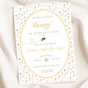 Peut inclure: Une invitation rectangulaire avec un motif floral et d'abeilles. Le texte "OUR LITTLE honey IS TURNING ONE!" est en écriture dorée. L'invitation comprend les détails d'une célébration de premier anniversaire, y compris la date, l'heure et le lieu.