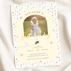 May include: A personalized invitation with a photo of a child in a white dress. The invitation features the text "Eleanor's first bee day" with a bee illustration and floral accents. The date, time, and location are also included.