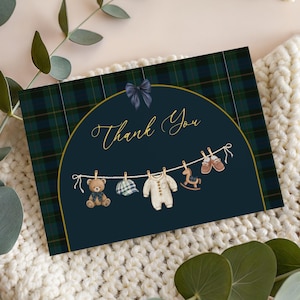 Puede incluir: Tarjeta de agradecimiento para baby shower con fondo de cuadros azul marino. La tarjeta presenta un diseño arqueado dorado con las palabras "Thank You" en escritura elegante. Una cuerda de ropa muestra ilustraciones temáticas de bebés.